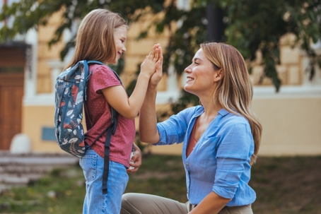 Mother kneeling outside giving her young daughter with a backpack a high five before school, showing support and reassurance.