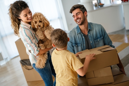 Smiling parents and young son carry moving boxes with their dog while relocating to a new Florida home.